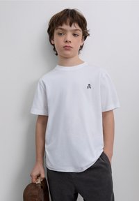 T-shirt en coton blanc avec un col rond et des manches courtes, arborant un petit logo de crâne noir sur la poitrine. Design simple, texture lisse.