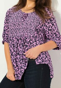 Blusa viola con motivo floreale nero, corpo arricciato, scollatura tonda con un laccio e maniche a tre quarti, abbinata a jeans scuri.
