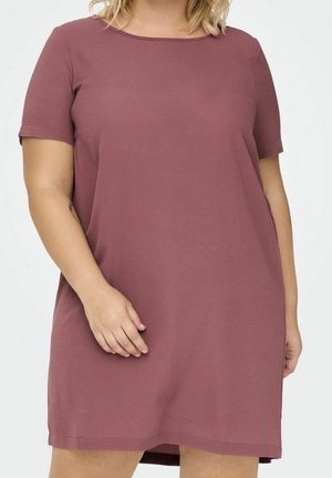 Kobieta ubrana w krótką sukienkę w kolorze mauve do kolan, stojąca z jedną ręką spoczywającą przy biodrze.
