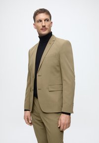 Lichtbruine kostuum met een slim fit ontwerp, een enkelbreasted blazer en vlakke broek, gecombineerd met een zwarte coltrui. Dure textuur.