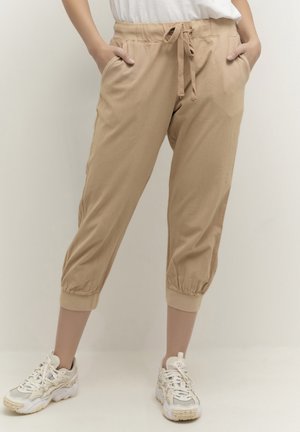 Person trägt beige kurz geschnittene Jogginghosen mit Kordelzug an der Taille und weiße klobige Sneaker, Hände in den Taschen, steht vor einem einfarbigen Hintergrund.