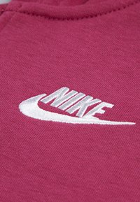 Bordeaux tkanina s textúrovaným povrchom; obsahuje bielo vyšívané logo Nike, ktoré je umiestnené v strede a zobrazuje zakrivený dizajn swoosh.
