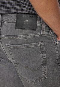 Grå denimjeans med en lærmerke, detaljer med søm på baklommen, og en lett falmet tekstur. Har standard design på baklommen.