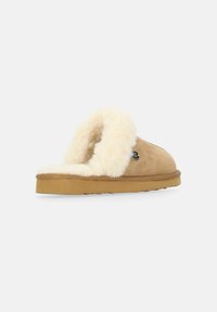 Tan mockasiner i mocka med en fluffig krämfärgad foder, öppen bakdesign, brun gummisula och en liten svart emblem på sidan.
