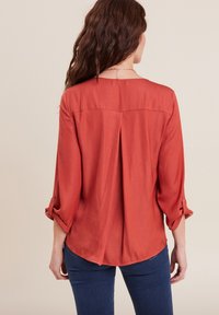 Blusa color ruggine con maniche a tre quarti, taglio dritto e design sul retro plissettato. Realizzata in tessuto leggero e morbido.