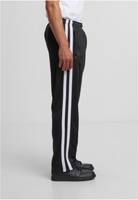 Crne sportske hlače s bijelim bočnim prugama, elastičnim pojasom, patentnim zatvaračima na rubovima i glatkom teksturom, prikazane kako se nose s crnim cipelama.