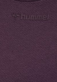 Tekstureret dyb lilla stof med hævet sort "hummel" logo og bie-symbol trykt nær øverste centrum.
