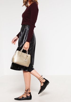 Vrouw die loopt, gekleed in een bordeauxrode top met lange mouwen, een zwarte plisséleren rok, zwarte veterschoenen en met een gouden handtas.