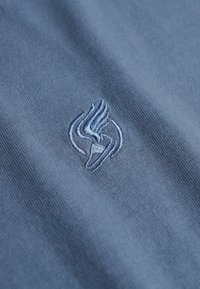 Tejido azul claro con una textura suave que presenta un logotipo bordado en un azul más oscuro, que representa un ala estilizada y un diseño circular.