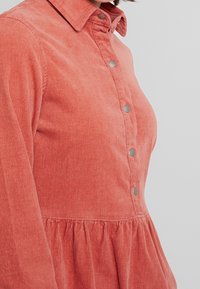 Korálové kordové šaty s předním zapínáním na knoflíky, špičatým límcem a nabíraným detailem sukně. Měkká textura a stříbrné knoflíky poskytují kontrast.