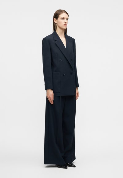 Γυναίκα με σκούρο, φαρδύ διπλόσκαρτο σακάκι με μεγάλες τσέπες και αντίστοιχο παντελόνι με φαρδιά μπατζάκια, όρθια μπροστά σε απλό φόντο.