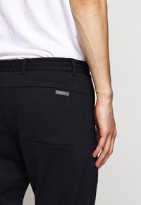 Donkere stretchstof broek met een gestructureerde tailleband, achterzak en een metalen accentlabel. Het ontwerp is eenvoudig en functioneel.