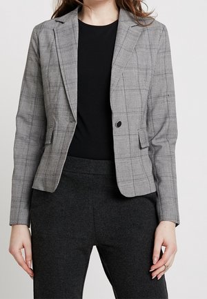 Kobieta nosząca dopasowaną szarą kratkę marynarkę na czarnej bluzce i ciemnoszarych spodniach, stojąca z rękami swobodnie opuszczonymi wzdłuż ciała.