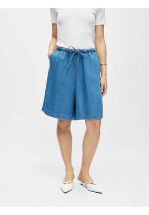 Femme portant un short en jean bleu mi-long avec taille élastique et lien, un haut blanc à manches courtes et des chaussures blanches pointues à talon bas.