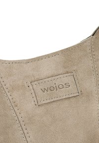 Gros plan sur une section de chaussure en daim beige avec un patch rectangulaire cousu affichant le nom de la marque "wojas".