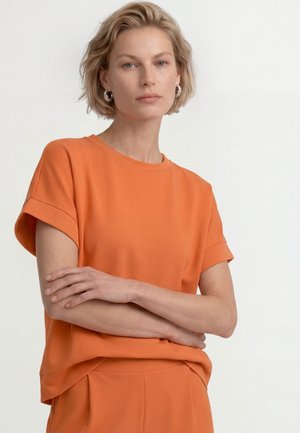 Lyhytkukkainen vaaleaverikköinen nainen, jolla on hopeiset renkaankorvakorut ja oranssi lyhythihainen paita sekä yhteen sopivat housut valkoisella taustalla.