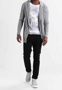 Grå langærmet cardigan over en grafisk hvid T-shirt, parret med sorte jeans og grå sneakers, med et moderne fit og afslappet design.