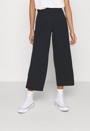 Pantalons larges noirs en tissu doux, dotés d'une taille haute, de poches latérales et d'une finition lisse, associés à des baskets montantes blanches.