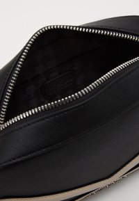 Černá zipová taška s texturovaným materiálem, lesklým kováním a textilním podšívkou se jemným vzorem. Mezi ozdobnými prvky se nachází i logo štítek.