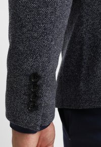 Manica di un blazer blu scuro texturizzato con quattro bottoni neri sopra un polso con cimosa e pantaloni scuri visibili.