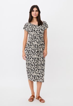 Femme debout portant une robe fleurie noire et blanche longueur genou avec des manches courtes et des sandales plates marron sur fond blanc.
