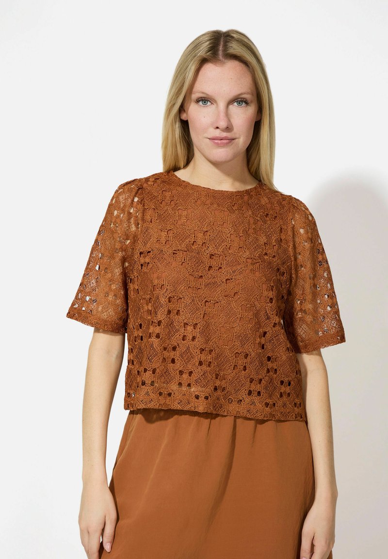 Blouse en dentelle marron avec un motif carré, des manches courtes et un col rond, portée sur une jupe marron coordonnée. Détail de tissu texturé visible.