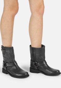 Botas de tobillo de cuero negro con punta redondeada, tacón bajo y una correa decorativa con hebilla de metal y tachuelas. Textura suave.
