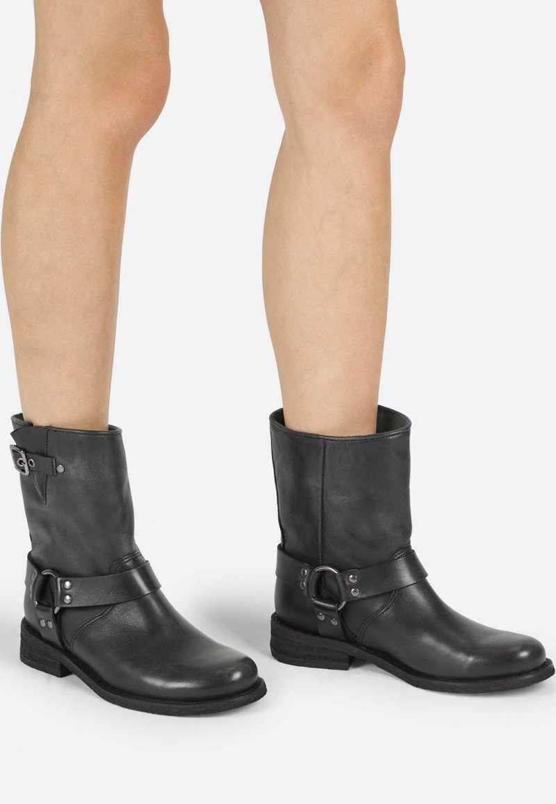 Botas de tobillo de cuero negro con punta redondeada, tacón bajo y una correa decorativa con hebilla de metal y tachuelas. Textura suave.