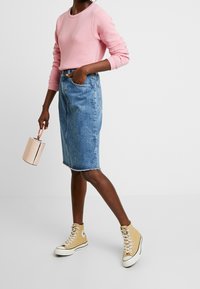 Ljusrosa långärmad tröja, högmidjade denimkjol och beige höga sneakers. Håller en liten cylindrisk väska med handtag.