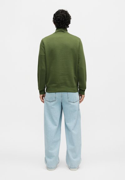 Άνδρας με κατσαρά μαλλιά που φοράει ελιάς πράσινο φούτερ και ανοιχτό μπλε τζιν με φαρδιά μπατζάκια, στέκεται με την πλάτη σε λευκό φόντο.