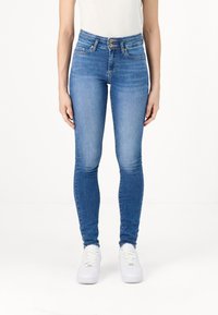 Jeans skinny de talle alto en denim azul. Cuenta con dos bolsillos frontales, cierre de doble botón y una textura suave. Combinados con zapatillas blancas.