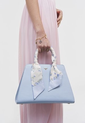 Sieviete rozā kleitā, tur rokās gaiši zilu trapecveida rokassomu ar ziedu šalli, kas ir sasieta ap somas rokturiem, un zelta bišu emblēmu.