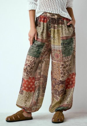 Moteris dėvi margus daugiabriaunius plataus kirpimo kelnes, baltą megztinį ir rudus zomšinius atvirus sandalų pirštus, stovinti su rankomis kišenėse.