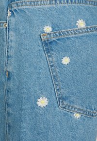 Hellblauer Denimstoff mit weißen, bestickten Gänseblümchen mit gelben Zentren, hauptsächlich im Bereich der hinteren Tasche dargestellt.