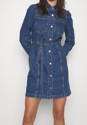 Woman wearing a knee-length blue denim dress with long sleeves, front buttons, chest pockets, and a tied belt at the waist.