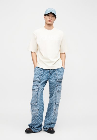 Homme portant un t-shirt ample de couleur beige clair, un jean cargo bleu à motifs, des chaussures noires et une casquette de baseball bleue, debout les mains dans les poches.