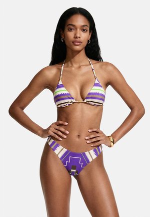Donna con capelli lunghi e neri che indossa un bikini viola, bianco e verde a fantasia, orecchini a cerchio d’oro e un braccialetto d’oro, in piedi con le mani sui fianchi.