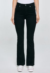 Nő fekete bootcut farmert és fehér tornacipőt visel, világosszürke háttér előtt áll, kezei lazán lógnak oldalán.