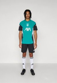 Športni dres v barvi modrozeleno in črni z logotipom Liverpoola, združen s črnimi kratkimi hlačami. Poudarjen z belimi črtami na rokavih, nogavicah in detajlih logotipa.