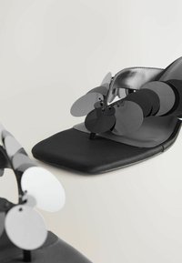 Svart platt sandal med fyrkantig tå, prydd med överlappande cirkulära detaljer i svart och vitt, tillverkad av ett glänsande material. Platt sula.