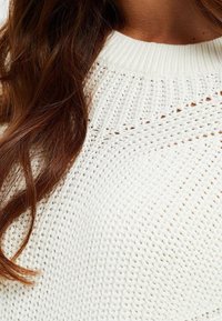 Stickad vit tröja med en texturerad, lös väv; har en ribbad halsringning och öppen vävdetaljering som visar en mjuk, avslappnad stil.