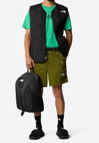 Black quilted vest over a green T-shirt, paired with olive shorts. Black backpack with mesh accents and black sandals, complemented by black socks.