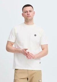 Krémové bavlněné tričko s kulatým výstřihem a krátkými rukávy, s malým černým geometrickým logem na prsou.