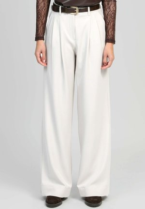 Femme portant un pantalon blanc taille haute à jambes larges avec des plis devant, une ceinture marron, un haut à manches longues en dentelle, et des chaussures marron, debout droite.