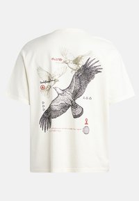 T-shirt couleur crème avec des illustrations d'oiseaux noirs et rouges, des formes géométriques et du texte dans différentes polices au dos. Manches courtes.