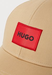 Casquette de baseball beige avec une visière courbée et un patch rectangulaire rouge portant le texte "HUGO" en noir. Tissu texturé avec des détails de couture visibles.
