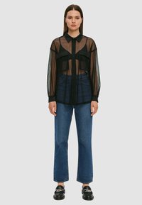 Camicia nera trasparente con bottoni e maniche lunghe, due tasche sul petto, indossata sopra jeans di denim blu a vita alta, abbinata a mocassini neri.