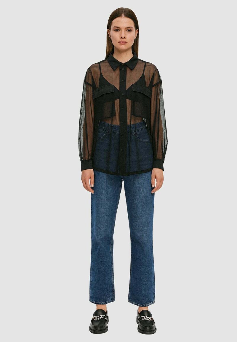 Camicia nera trasparente con bottoni e maniche lunghe, due tasche sul petto, indossata sopra jeans di denim blu a vita alta, abbinata a mocassini neri.