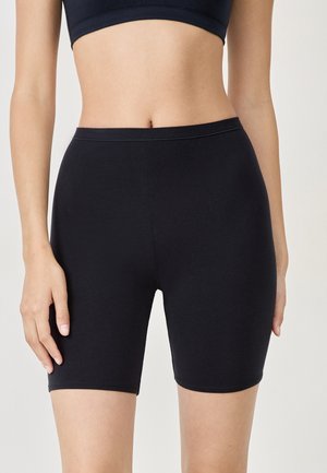 Frau trägt hoch taillierte schwarze Biker-Shorts und ein passendes kurzes Top, steht vor einem schlichten hellen Hintergrund.