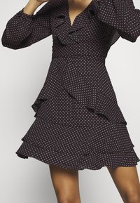 Femme portant une robe noire à pois roses, avec des volants superposés sur la jupe et un décolleté en V avec des manches longues.